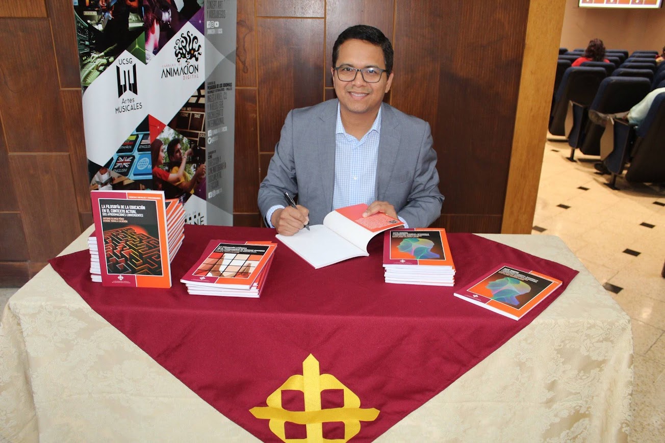 Byrone Tomalá sonriente, firmando un libro en una mesa con publicaciones